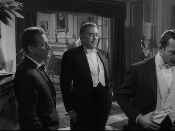 Movie still from “The Exterminating Angel” (1962), directed by Luis Buñuel – A black and white photo of three men in tuxedos in front of a fireplace; Medium shot, Over the shoulder angle