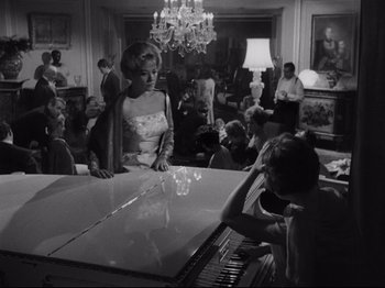 Movie still from “The Exterminating Angel” (1962), directed by Luis Buñuel – A woman standing next to a baby grand piano; Wide shot, High angle