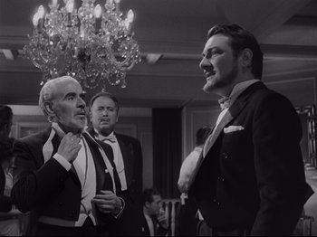 Movie still from “The Exterminating Angel” (1962), directed by Luis Buñuel – A group of men standing next to each other in front of a chandelier; Medium shot, Over the shoulder angle