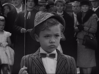 Movie still from “The Exterminating Angel” (1962), directed by Luis Buñuel – A young boy in a suit and a bow tie in front of a group of people; Medium shot, High angle