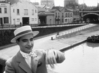 Movie still from “The Fiancés of the Bridge Mac Donald” (1961), directed by Agnès Varda – A man in a suit and a hat pointing to the side; Medium shot, Over the shoulder angle