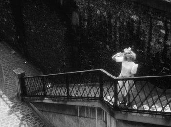 Movie still from “The Fiancés of the Bridge Mac Donald” (1961), directed by Agnès Varda – A black and white photo of a woman standing on a railing; Wide shot, High angle