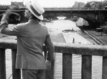 Movie still from “The Fiancés of the Bridge Mac Donald” (1961), directed by Agnès Varda – A man in a suit and hat looking over a bridge; Wide shot, High angle