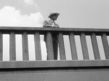 Movie still from “The Fiancés of the Bridge Mac Donald” (1961), directed by Agnès Varda – A black and white photo of a man standing on a bridge; Wide shot, Low angle