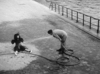 Movie still from “The Fiancés of the Bridge Mac Donald” (1961), directed by Agnès Varda – A man and a woman are playing with a water hose; Wide shot, High angle
