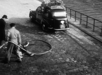 Movie still from “The Fiancés of the Bridge Mac Donald” (1961), directed by Agnès Varda – An old photo of a man cleaning the sidewalk with a bicycle; Wide shot, High angle