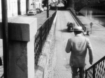 Movie still from “The Fiancés of the Bridge Mac Donald” (1961), directed by Agnès Varda – A man walking down a street with a motorcycle behind him; Wide shot, High angle
