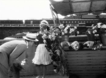 Movie still from “The Fiancés of the Bridge Mac Donald” (1961), directed by Agnès Varda – An old photo of a woman standing next to a bunch of flowers; Wide shot, Low angle