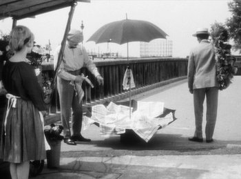 Movie still from “The Fiancés of the Bridge Mac Donald” (1961), directed by Agnès Varda – An old black and white photo of a man and a woman standing next to an umbrella; Wide shot, High angle