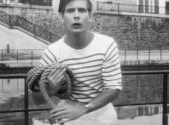 Movie still from “The Fiancés of the Bridge Mac Donald” (1961), directed by Agnès Varda – A man holding a tennis racquet on a tennis court; Medium shot, High angle