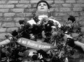 Movie still from “The Fiancés of the Bridge Mac Donald” (1961), directed by Agnès Varda – A man standing next to a brick wall holding a bunch of flowers; Medium shot, Low angle