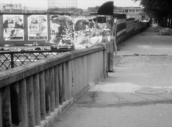 Movie still from “The Fiancés of the Bridge Mac Donald” (1961), directed by Agnès Varda – A man standing next to a railing on a sidewalk; Wide shot, Over the shoulder angle
