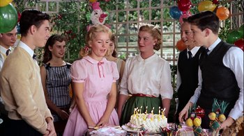 Movie still from “The Five Pennies” (1959), directed by Melville Shavelson – A group of young people standing around a cake; Medium shot, Over the shoulder angle