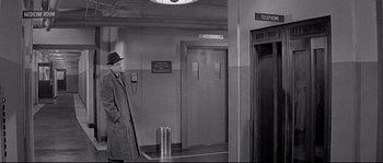 Movie still from “The Fortune Cookie” (1966), directed by Billy Wilder – A man in a hat and a long coat standing in an elevator; Wide shot, High angle