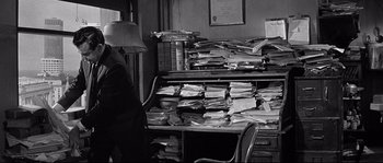 Movie still from “The Fortune Cookie” (1966), directed by Billy Wilder – A person standing in front of a desk with papers on it; Medium shot, High angle
