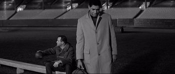 Movie still from “The Fortune Cookie” (1966), directed by Billy Wilder – A man in a trench coat is standing next to another man in a field; Medium shot, Low angle