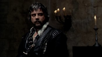 Movie still from “The Four Musketeers: Milady's Revenge” (1974), directed by Richard Lester – A man in a black and white outfit standing in front of candles; Medium shot, Over the shoulder angle