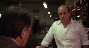 Movie still from “The Friends of Eddie Coyle” (1973), directed by Peter Yates – A man with a bald head talking to another man in front of a counter; Medium shot, Over the shoulder angle