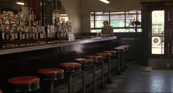 Movie still from “The Friends of Eddie Coyle” (1973), directed by Peter Yates – A man sitting at a counter in front of red stools in a restaurant; Wide shot, Over the shoulder angle
