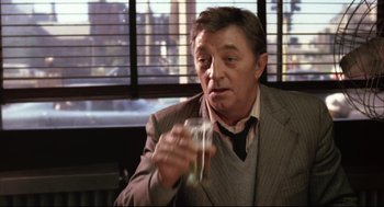 Movie still from “The Friends of Eddie Coyle” (1973), directed by Peter Yates – A man sitting in a chair holding a glass of water; Medium shot, Over the shoulder angle
