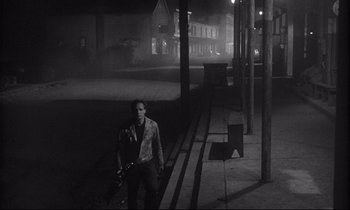 Movie still from “The Fugitive Kind” (1960), directed by Sidney Lumet – A man standing on the sidewalk holding a gun in his hand; Wide shot, High angle