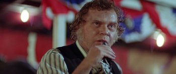 Movie still from “The Funhouse” (1981), directed by Tobe Hooper – A man with curly hair and a tie is sitting in front of a flag; Close Up shot, Low angle