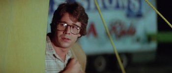 Movie still from “The Funhouse” (1981), directed by Tobe Hooper – A man with glasses is sitting in front of a sign; Close Up shot, Over the shoulder angle