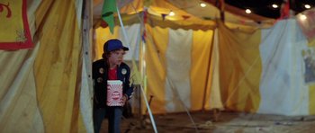 Movie still from “The Funhouse” (1981), directed by Tobe Hooper – A young boy standing in front of a tent holding a box; Medium shot, Over the shoulder angle