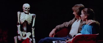 Movie still from “The Funhouse” (1981), directed by Tobe Hooper – Two people sitting on a red chair in front of a skeleton; Medium shot, Over the shoulder angle