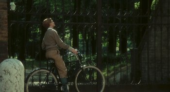 Movie still from “The Garden of the Finzi-Continis” (1970), directed by Vittorio De Sica – A person riding a bike near a metal fence; Wide shot, High angle