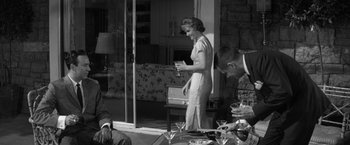 Movie still from “The Gazebo” (1959), directed by George Marshall – A woman standing in front of a table holding a cell phone; Medium shot, Over the shoulder angle