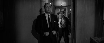 Movie still from “The Gazebo” (1959), directed by George Marshall – A man in a suit standing next to another man in a hat; Medium shot, Over the shoulder angle