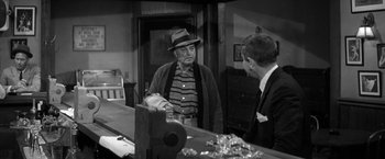 Movie still from “The Gazebo” (1959), directed by George Marshall – A man wearing a hat and a striped cardigan talking to another man in front of a counter; Medium shot, Over the shoulder angle