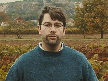 Movie still from “The Gleaners & I” (2000), directed by Agnès Varda – A man wearing glasses and a sweater standing in front of a field; Close Up shot, Low angle