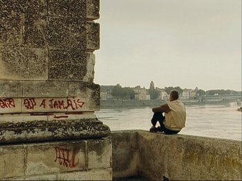 Movie still from “The Gleaners & I” (2000), directed by Agnès Varda – A man sitting on the side of a building next to a body of water; Extreme Wide shot, High angle