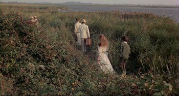 Movie still from “The Go-Between” (1971), directed by Joseph Losey – A group of people walking through tall grass near a body of water; Wide shot, High angle