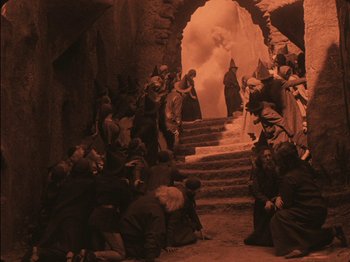 Movie still from “The Golem” (1920), directed by Paul Wegener – A group of people gathered in a cave with smoke billowing out of the entrance; Wide shot, High angle