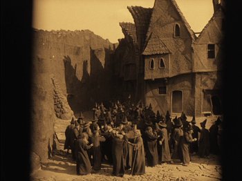 Movie still from “The Golem” (1920), directed by Paul Wegener – A group of people dressed as witches gathered outside of a house; Extreme Wide shot, High angle