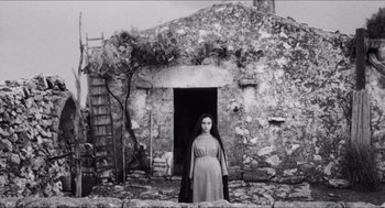 Movie still from “The Gospel According to St. Matthew” (1964), directed by Pier Paolo Pasolini – A woman standing in front of an old stone building; Wide shot, Low angle