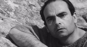 Movie still from “The Gospel According to St. Matthew” (1964), directed by Pier Paolo Pasolini – Black and white photograph of a man leaning against a rock wall; Close Up shot, Low angle