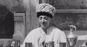 Movie still from “The Gospel According to St. Matthew” (1964), directed by Pier Paolo Pasolini – An older man wearing a turban and smiling for the camera; Medium shot, Low angle