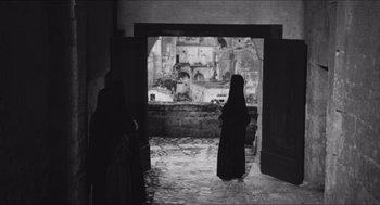 Movie still from “The Gospel According to St. Matthew” (1964), directed by Pier Paolo Pasolini – A woman in a black dress standing in front of an open door; Wide shot, Low angle