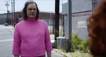 Movie still from “The Greasy Strangler” (2016), directed by Jim Hosking – An older woman wearing a pink shirt standing on the side of the road; Medium shot, Over the shoulder angle