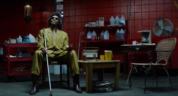 Movie still from “The Greasy Strangler” (2016), directed by Jim Hosking – A man in a yellow suit sitting on a chair in a room; Wide shot, Low angle