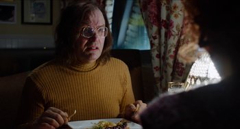 Movie still from “The Greasy Strangler” (2016), directed by Jim Hosking – An older woman sitting at a table with a plate of food in front of her; Medium shot, Over the shoulder angle