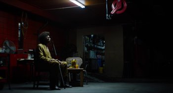 Movie still from “The Greasy Strangler” (2016), directed by Jim Hosking – A man sitting on a chair in a room; Wide shot, Low angle