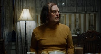 Movie still from “The Greasy Strangler” (2016), directed by Jim Hosking – A man with long hair and glasses sitting at a dinner table; Medium shot, Over the shoulder angle