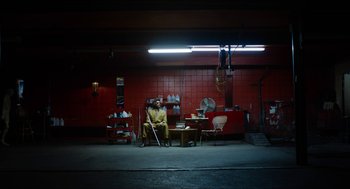 Movie still from “The Greasy Strangler” (2016), directed by Jim Hosking – A man sitting on a chair in a garage; Extreme Wide shot, Low angle