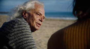 Movie still from “The Greasy Strangler” (2016), directed by Jim Hosking – An older man sitting on the beach looking into the distance; Close Up shot, Over the shoulder angle