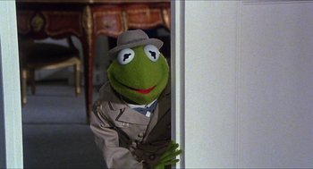 Movie still from “The Great Muppet Caper” (1981), directed by Jim Henson – A man dressed as kermit the frog peeks out from behind a wall; Medium shot, Over the shoulder angle